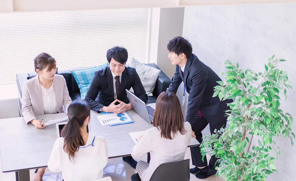 オフィスで4人のビジネスパーソンが資料を囲みながらミーティングをしている様子